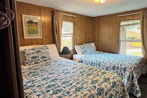 A room with two beds, wooden walls, a framed picture, and a window with curtains.