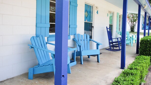 Un edificio con fachada blanca y detalles azules, incluyendo contraventanas y columnas. Hay zonas de estar al aire libre con sillas y una mesa azules.