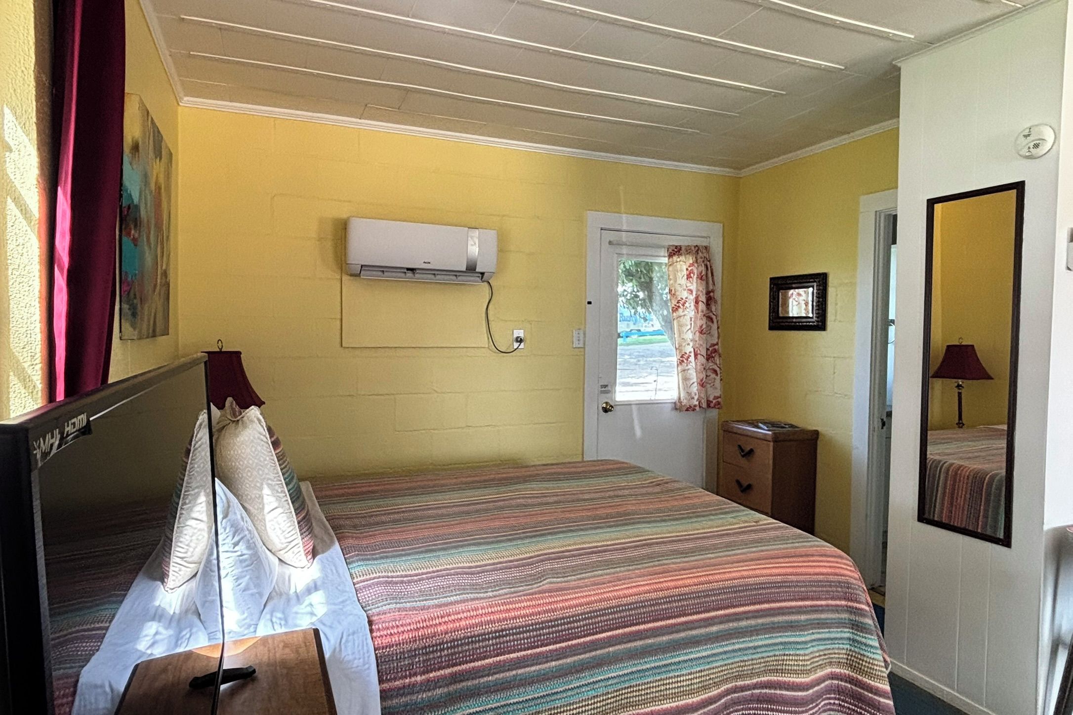 A room with a bed, a bedside table, a mirror, a door, and an air conditioning unit.