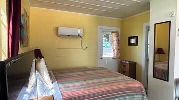A room with a bed, a bedside table, a mirror, a door, and an air conditioning unit.