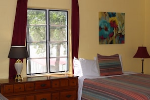 A room with a bed, a dresser, a window with curtains, a painting on the wall, and a lamp. - Cabin Steps Away from lake, Long‑Term Stay w/ WiFi & Pool (Kingsland)