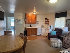 A compact living space with a wooden dining table, a sofa, a small kitchen with a stove and refrigerator, and a window with blinds. - Lakefront Suite - 15 (Kingsland)