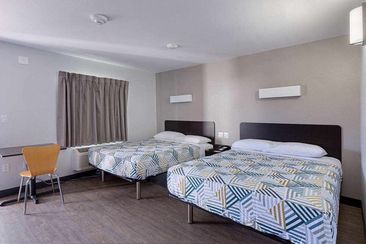 A hotel room with two single beds, a desk, a chair, and a window with curtains.