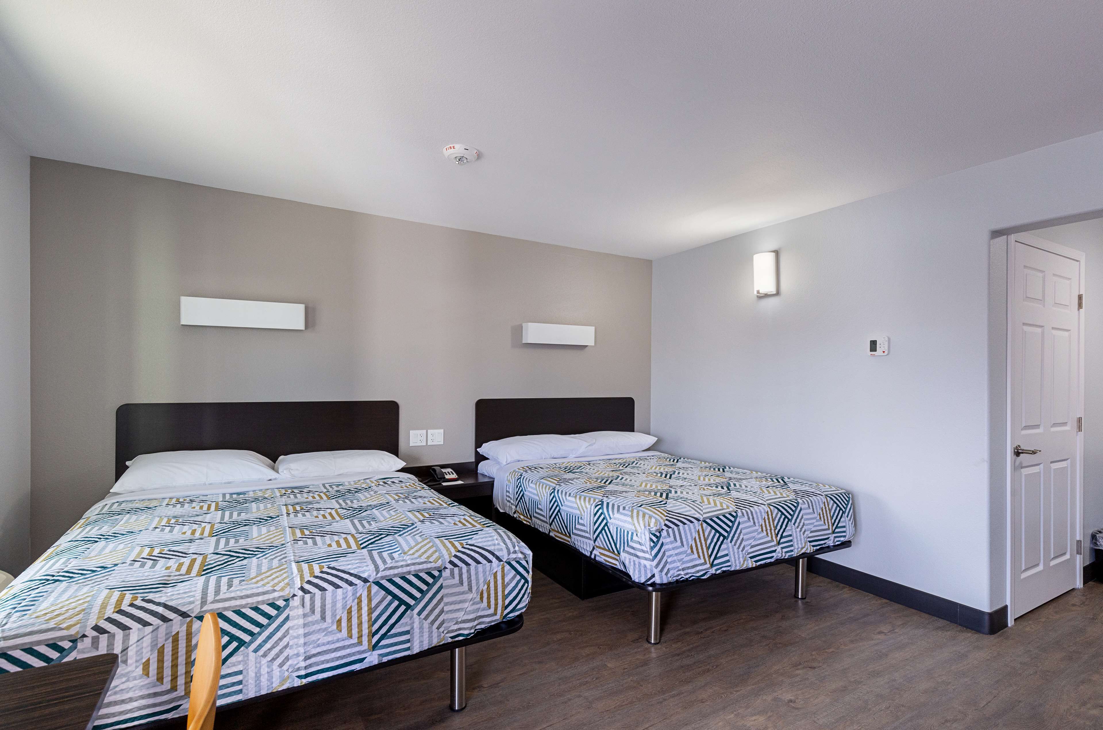 A hotel room with two single beds, a nightstand, a bedside table, a wall-mounted light, and a door.