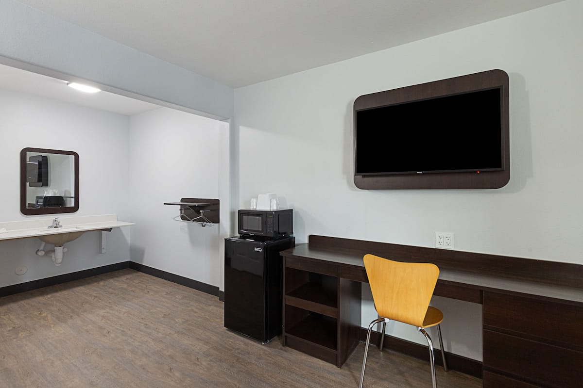 A hotel room with a flat-screen TV mounted on the wall, a sink with a mirror, a compact refrigerator, and a wooden desk with a chair.