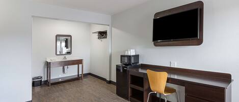 Une chambre d’hôtel avec un bureau en bois, une chaise, un téléviseur fixé au mur, un miroir et une petite étagère sur laquelle se trouvent un micro-ondes et une machine à café.