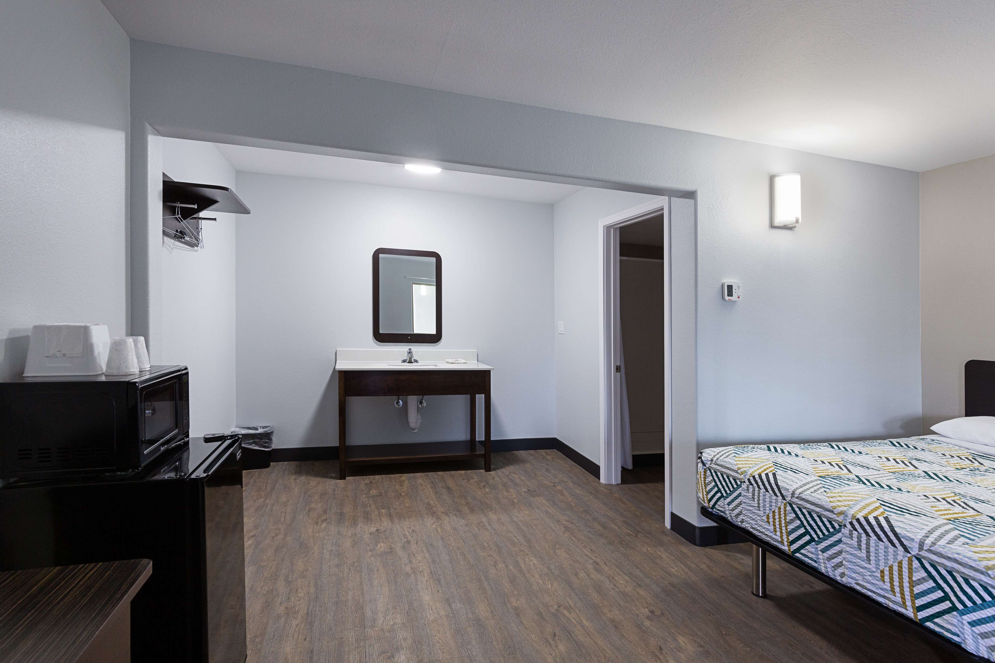 Habitación de hotel con piso de madera, cama con cubrecama estampado, lavamanos blanco con espejo arriba, una mesita con microondas y una puerta que da a otra habitación.