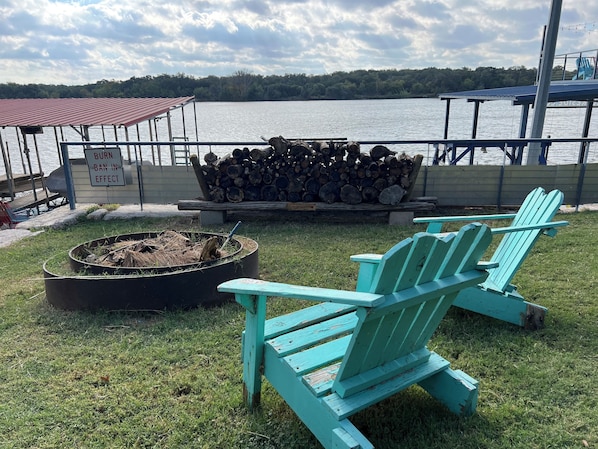 A lakeside deck with a fire pit, stacked firewood, and two turquoise Adirondack chairs.
