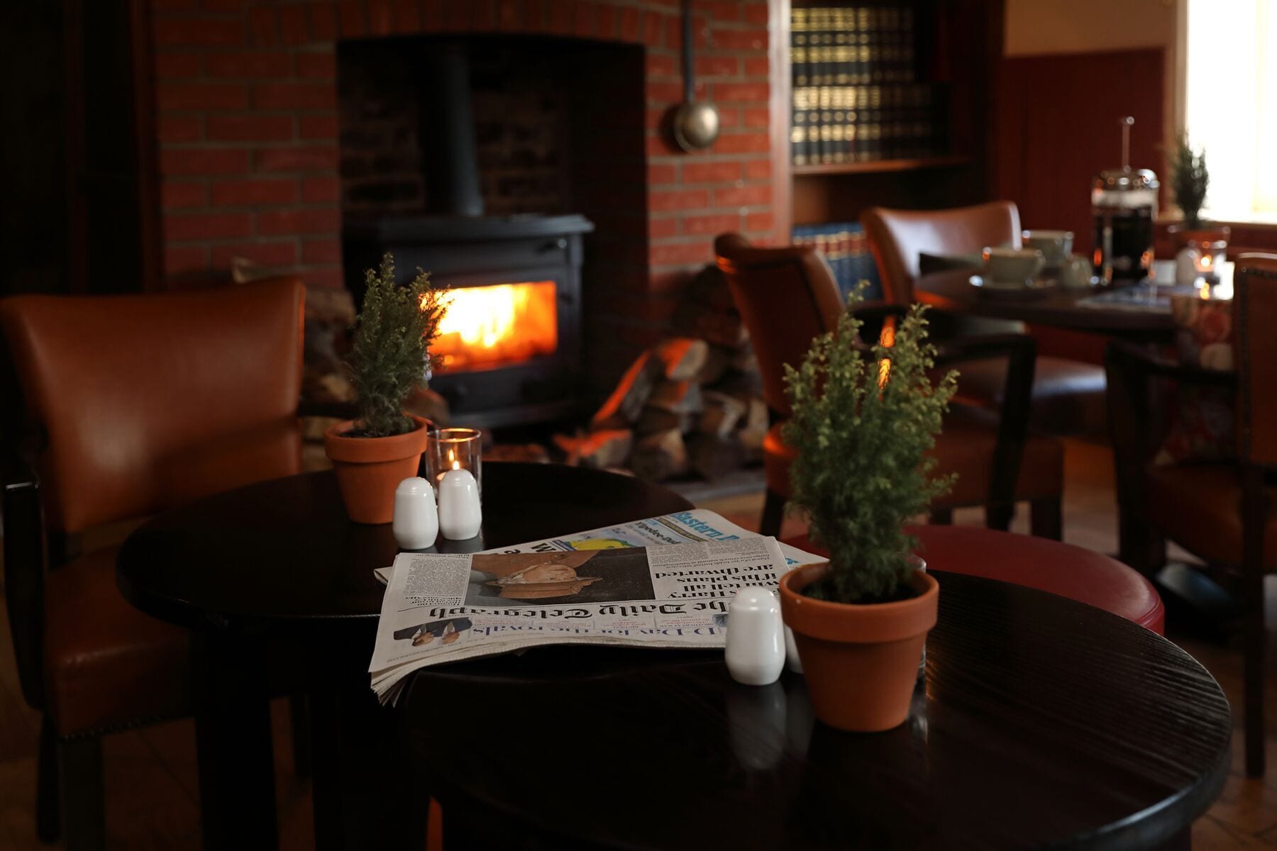 Un ambiente acogedor con chimenea de ladrillo, dos plantas en macetas, saleros y pimenteros, y un periódico sobre una mesa redonda.