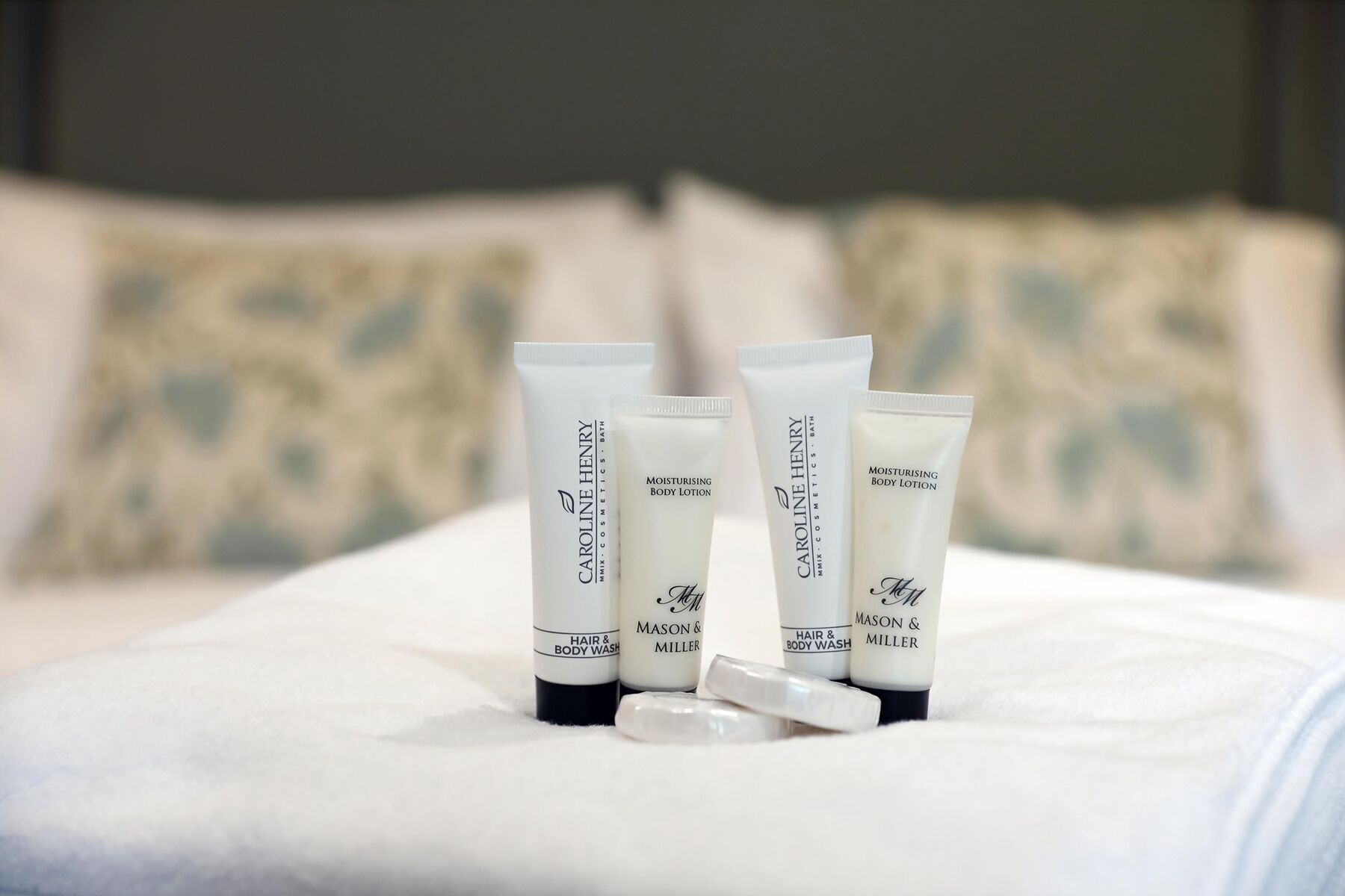 Four tubes of Caroline Henry hair and body wash on a bed with pillows.