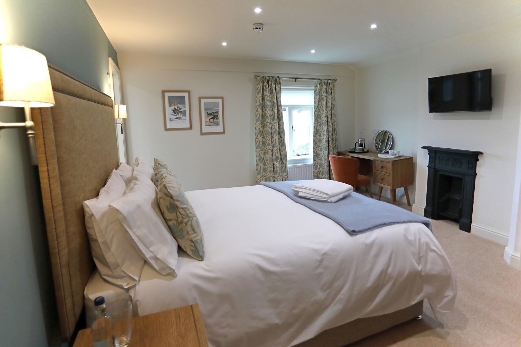 A neatly arranged bedroom with a bed, bedside table, lamp, framed pictures, a window with curtains, a desk with a mirror, and a fireplace.