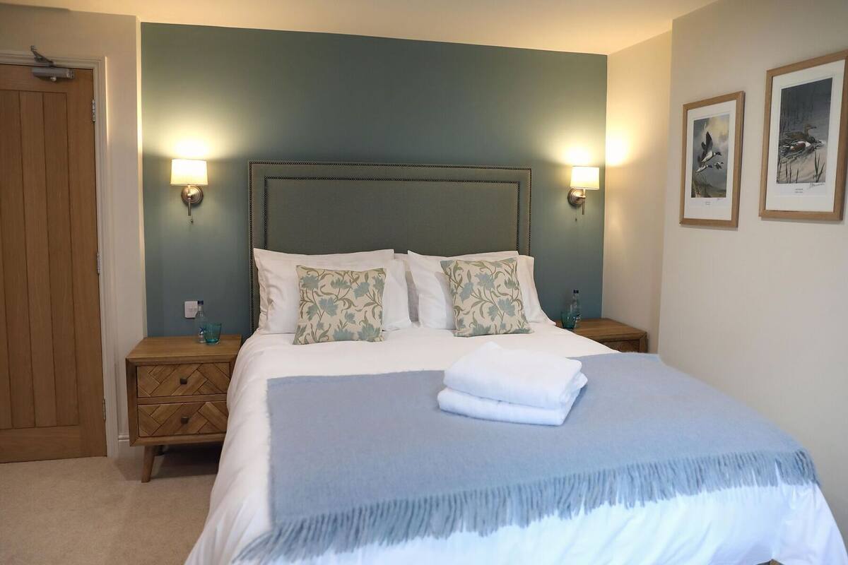 A neatly made bed with a blue blanket and two pillows, flanked by two bedside tables with lamps, in a room with a wooden door and a light blue wall.