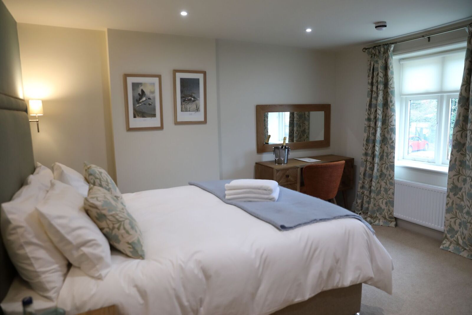 Une chambre d’hôtel avec un lit, un bureau et une chaise, un miroir et deux tableaux encadrés au mur.