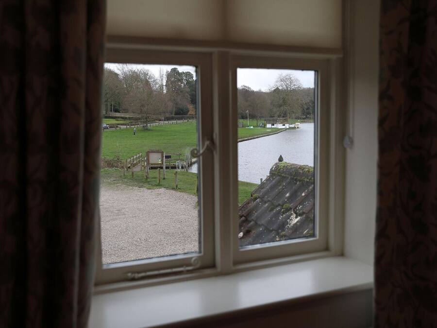Vista a través de una ventana de madera, que da a una zona verde con una construcción de madera pequeña y un cuerpo de agua.