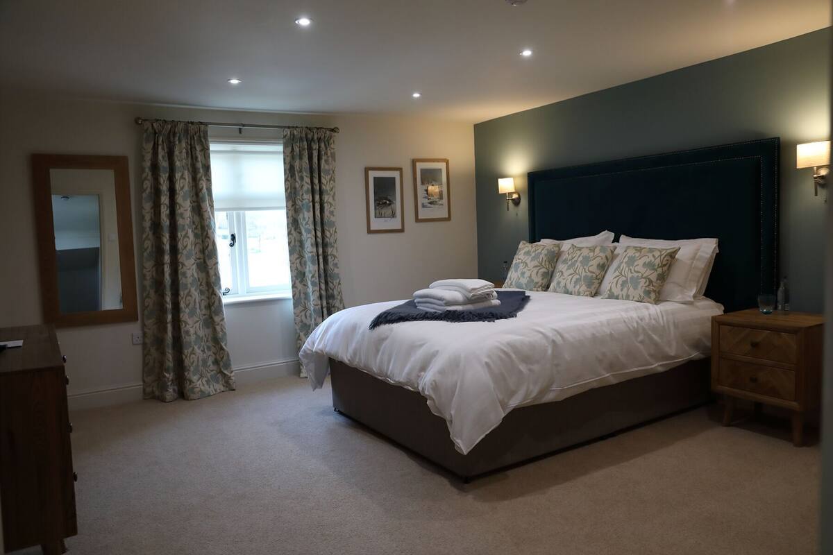 A bedroom with a large bed, a nightstand, a dresser, a window with curtains, and wall-mounted lights.