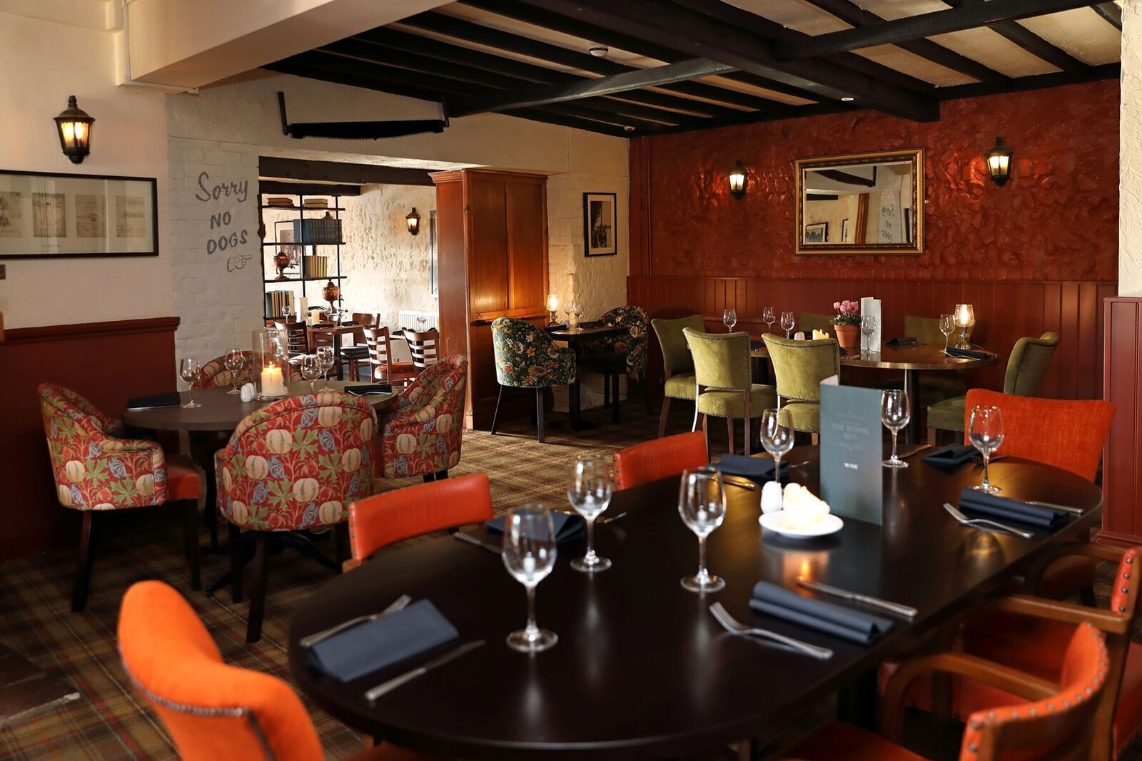 A restaurant with a mix of wooden and upholstered chairs, a long table set with wine glasses, and a 'Sorry No Dogs' sign on the wall.