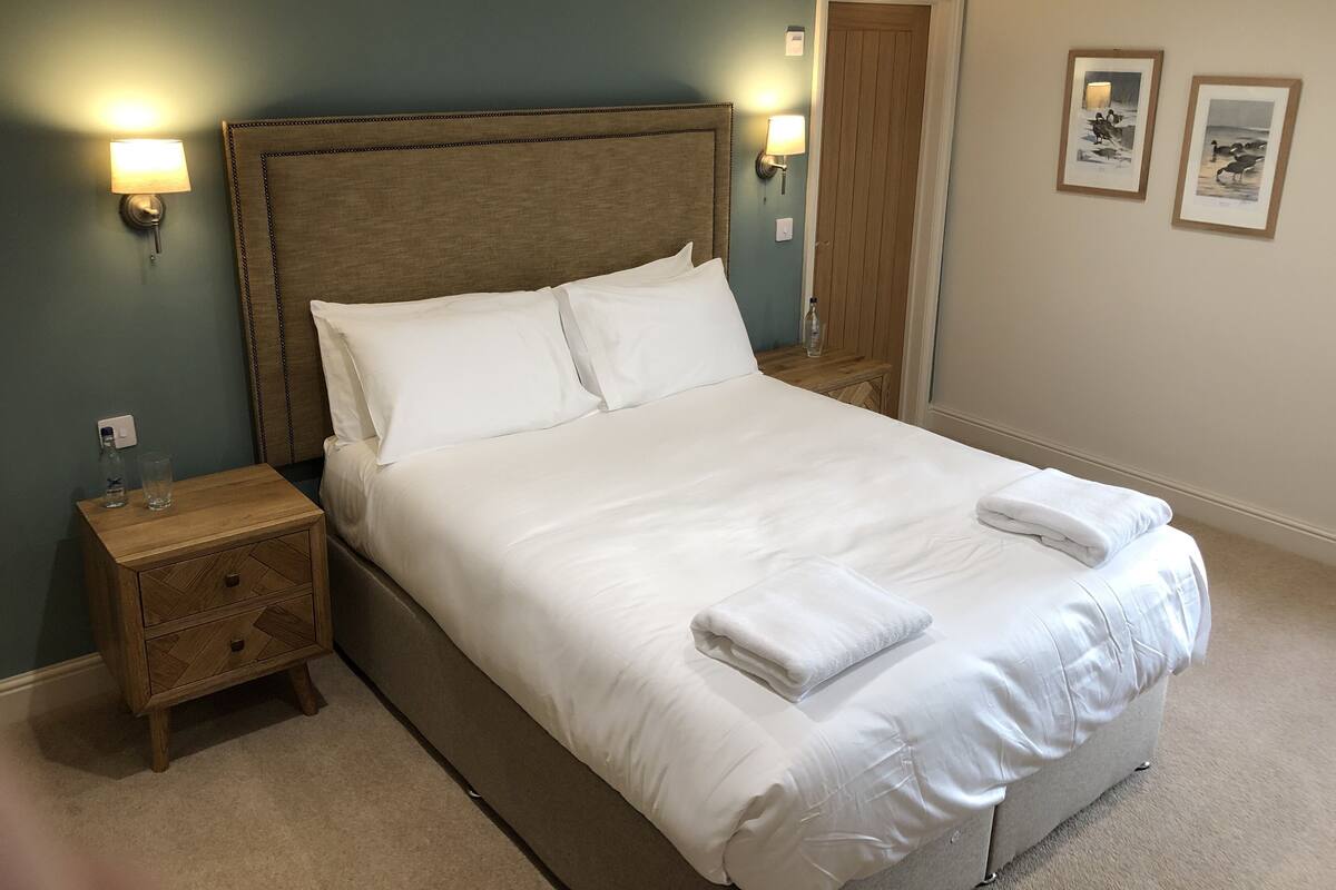 A neatly made bed with white linens and pillows, a wooden nightstand with a glass and bottle, and two wall-mounted lamps.