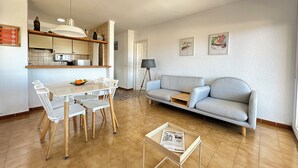A modern living room with a grey sofa, white dining table, and chairs. There is a kitchen area with a microwave and a lamp. - Agi Horitzo 11 Apartamento for rent in Roses Girona. (Roses)