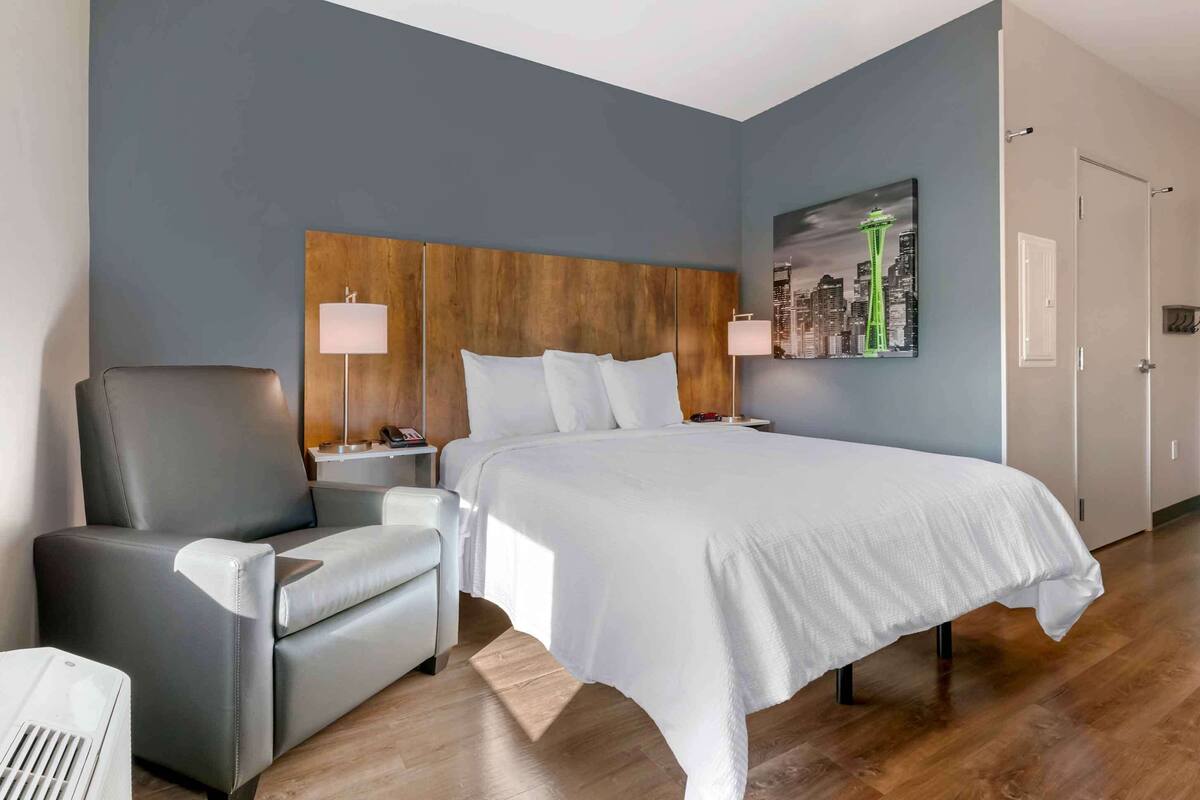 A hotel room with a large bed, a grey armchair, a bedside table with a lamp, and a cityscape painting on the wall.