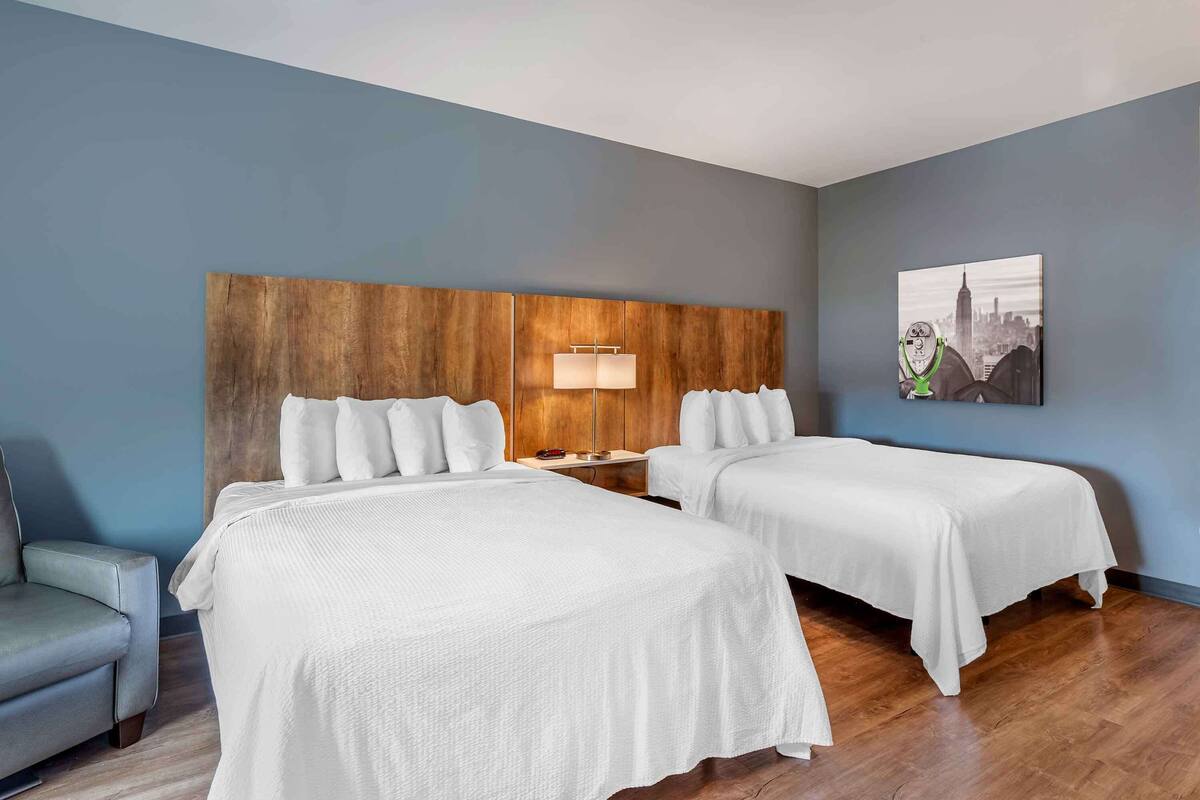 A hotel room with two beds, a wooden headboard, a nightstand with a lamp, a gray armchair, and a cityscape painting on the wall.
