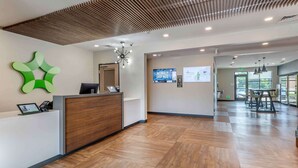 A reception area with a wooden reception desk, a green abstract wall art, a digital display, and a large window providing natural light. - Extended Stay America Suites Colonial Heights Fort Lee (Colonial Heights)