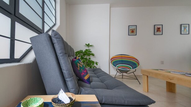 Ein modernes Wohnzimmer mit einem grauen Sofa, einem farbenfrohen Webstuhl-Sessel, einer Holzbank und einem kleinen Tisch mit dekorativen Schalen.