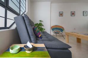 Ein modernes Wohnzimmer mit einem grauen Sofa, einem farbenfrohen Webstuhl-Sessel, einer Holzbank und einem kleinen Tisch mit dekorativen Schalen.