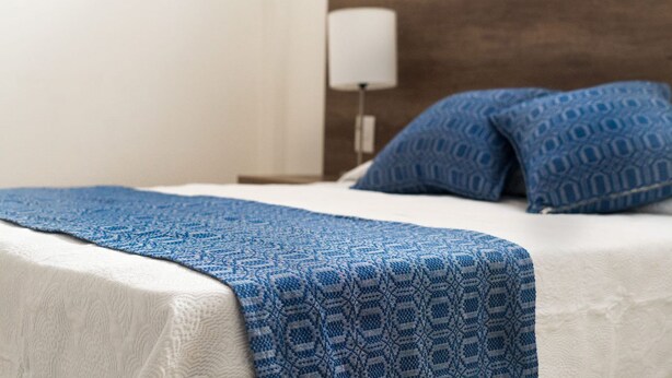 A neatly made bed with a blue patterned blanket and pillows, a white wall, and a wooden headboard. - JUUB at THE SOUL OF OAXACA (Oaxaca)