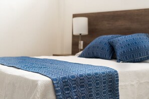 A neatly made bed with a blue patterned blanket and pillows, a white wall, and a wooden headboard.