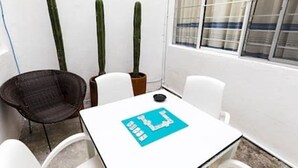 A small, white table with a blue placemat, two potted cacti, and a wicker chair in a corner of a room with white walls and a window with blinds. - Oaxaca at your doorstep (Oaxaca)