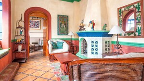 Quarto tradicional com chão em azulejos, lareira com mantelha azul, sofá branco com almofadas estampadas e um banco de madeira.