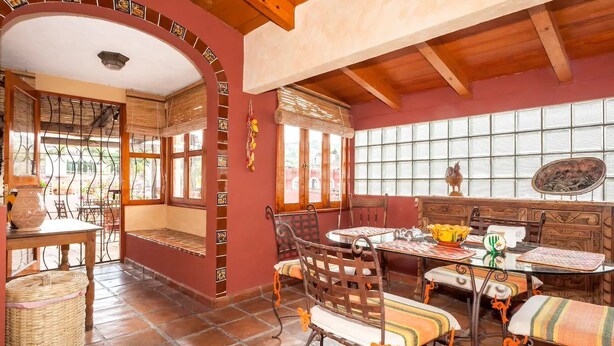 Un coin repas comprenant un banc en bois, une table et des chaises. Il y a une fenêtre avec des stores et une décoration murale.