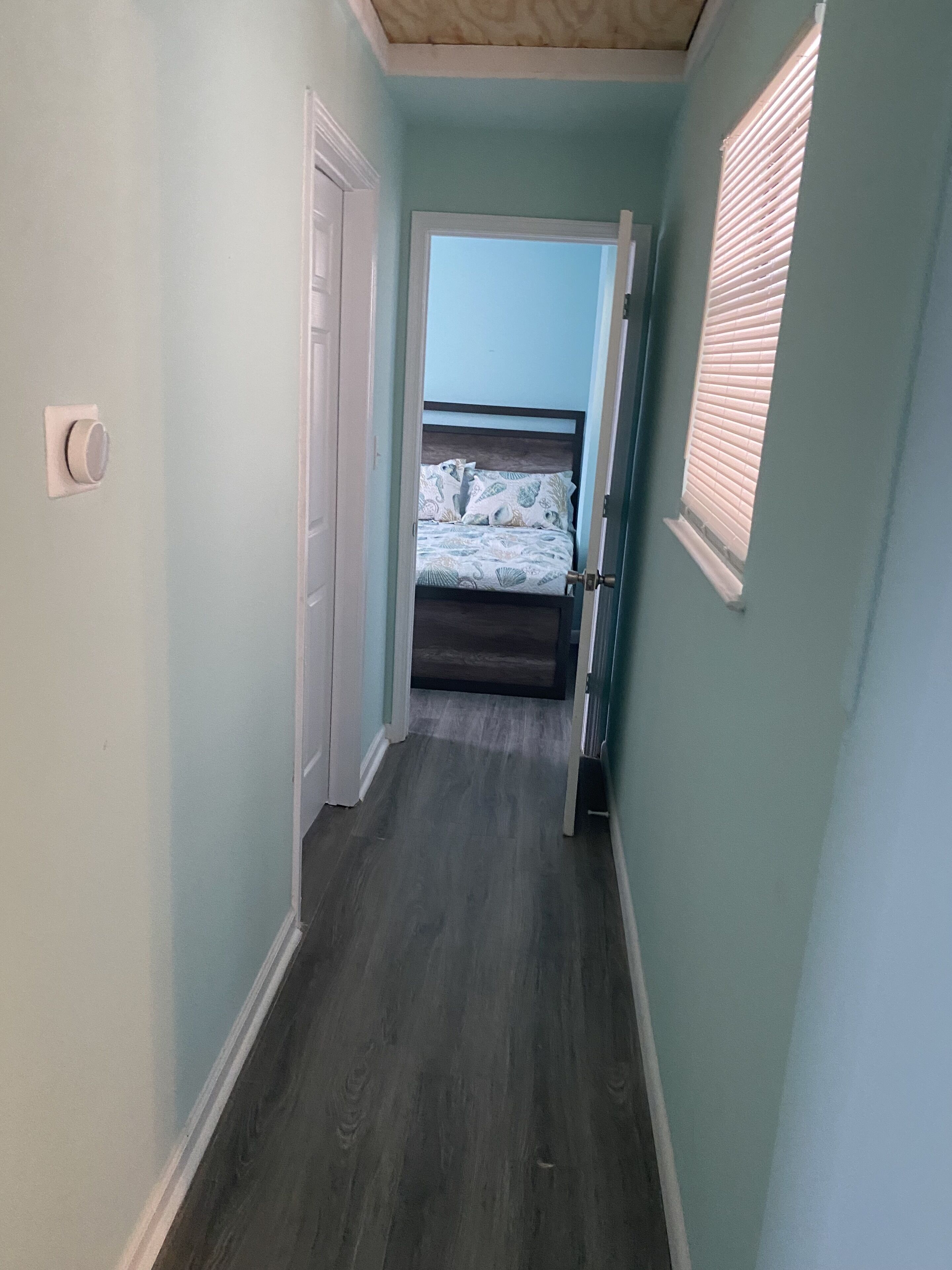 Um corredor estreito com paredes azul claro e soalho de madeira escura, que dá para um quarto com cama e janela com persianas.