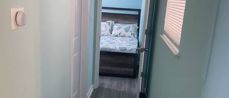 Um corredor estreito com paredes azul claro e soalho de madeira escura, que dá para um quarto com cama e janela com persianas.