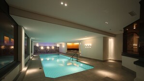 Una piscina amplia en el interior con suelo de baldosas, rodeada de paredes con luminarias empotradas y una pared decorativa de piedra.
