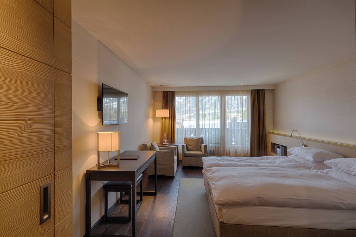 Quarto de hotel com cama, escrivaninha, cadeira, televisão e janela com cortinas.