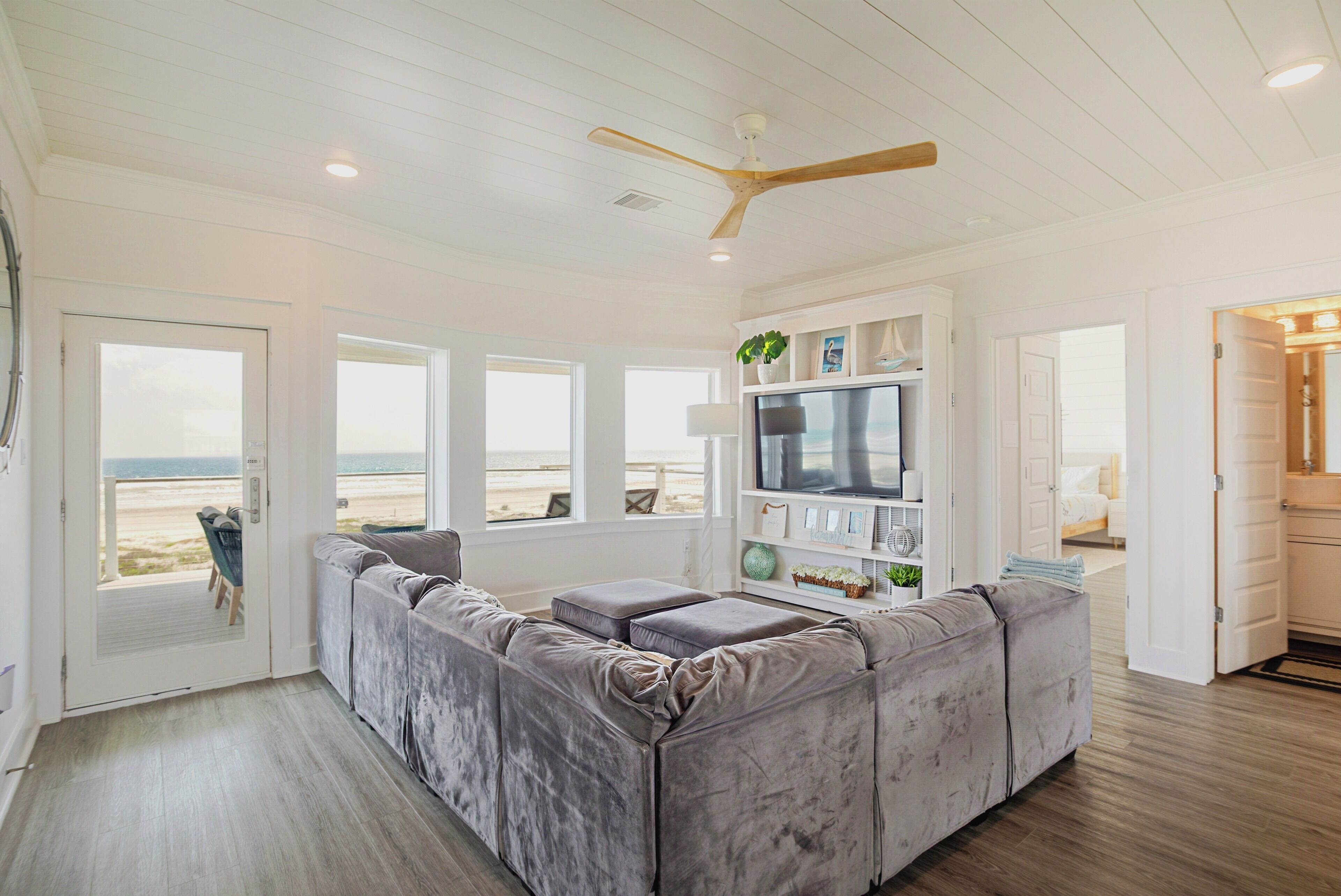 Un salon doté d’un grand canapé d’angle gris, d’un ventilateur de plafond, d’un téléviseur posé sur une étagère blanche et d’une porte donnant sur une vue sur la plage.
