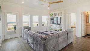 A living room with a large grey sectional sofa, a ceiling fan, a TV on a white shelf, and a door leading to a beach view.