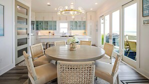A dining area with a round table, wicker chairs, a chandelier, and a view of the ocean through large windows.