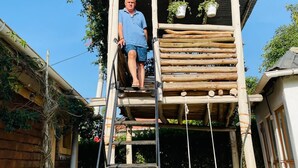 A man standing on a wooden ladder leading to a wooden platform with hanging planters.