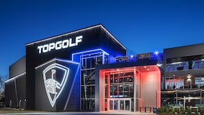 Ein modernes Gebäude mit auffälligem TOPGOLF-Schild, großem Glaseingang und einer gut beleuchteten Fassade.