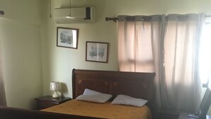 Ein Schlafzimmer mit einem hölzernen Bett, einem Nachttisch, einer Lampe, einem Schreibtisch mit einem Computer und zwei gerahmten Bildern an der Wand.