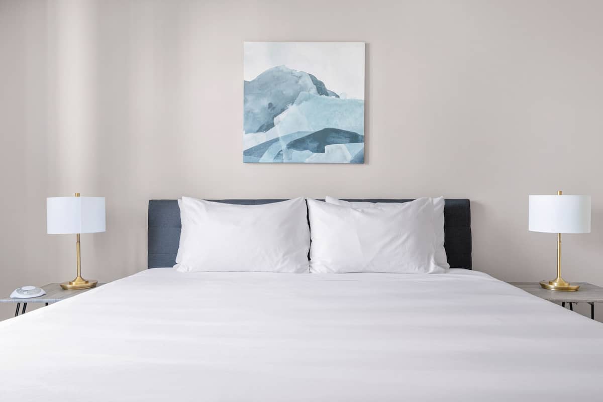 Une chambre d’hôtel avec un grand lit, du linge de lit blanc, deux tables de chevet avec des lampes et un tableau représentant une montagne, accroché au mur.