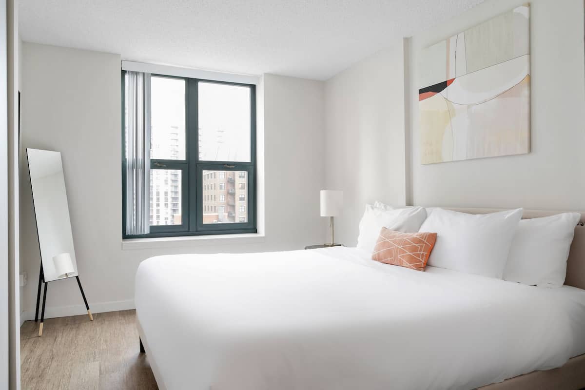 Une chambre d’hôtel avec un grand lit, une lampe sur pied, un tableau au mur et une fenêtre donnant sur des immeubles.
