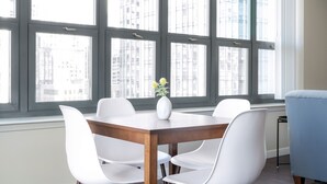 Un comedor con una mesa de madera y sillas blancas, ventanas amplias con persianas y un jarrón con flores sobre la mesa.