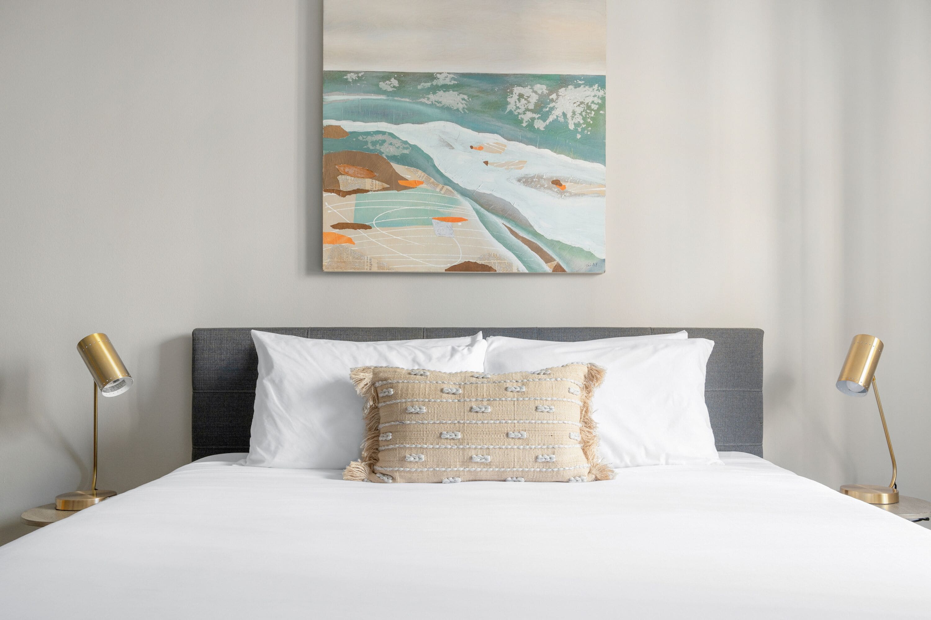 A neatly made bed with a patterned pillow, a wall-mounted painting, and two bedside lamps.