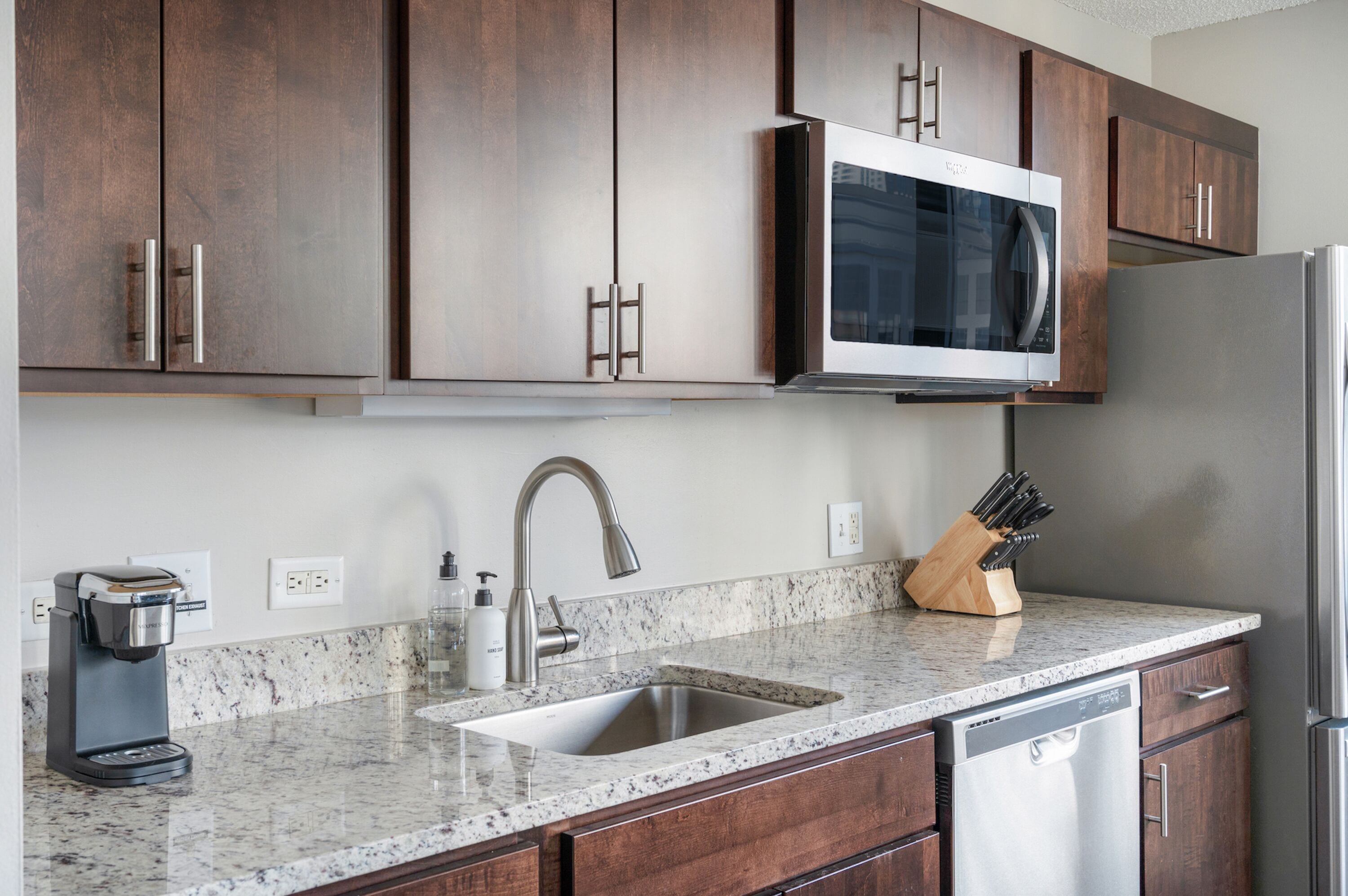 A kitchen with dark wood cabinets, a stainless steel refrigerator, a built-in microwave, a coffee maker, and a knife block.
