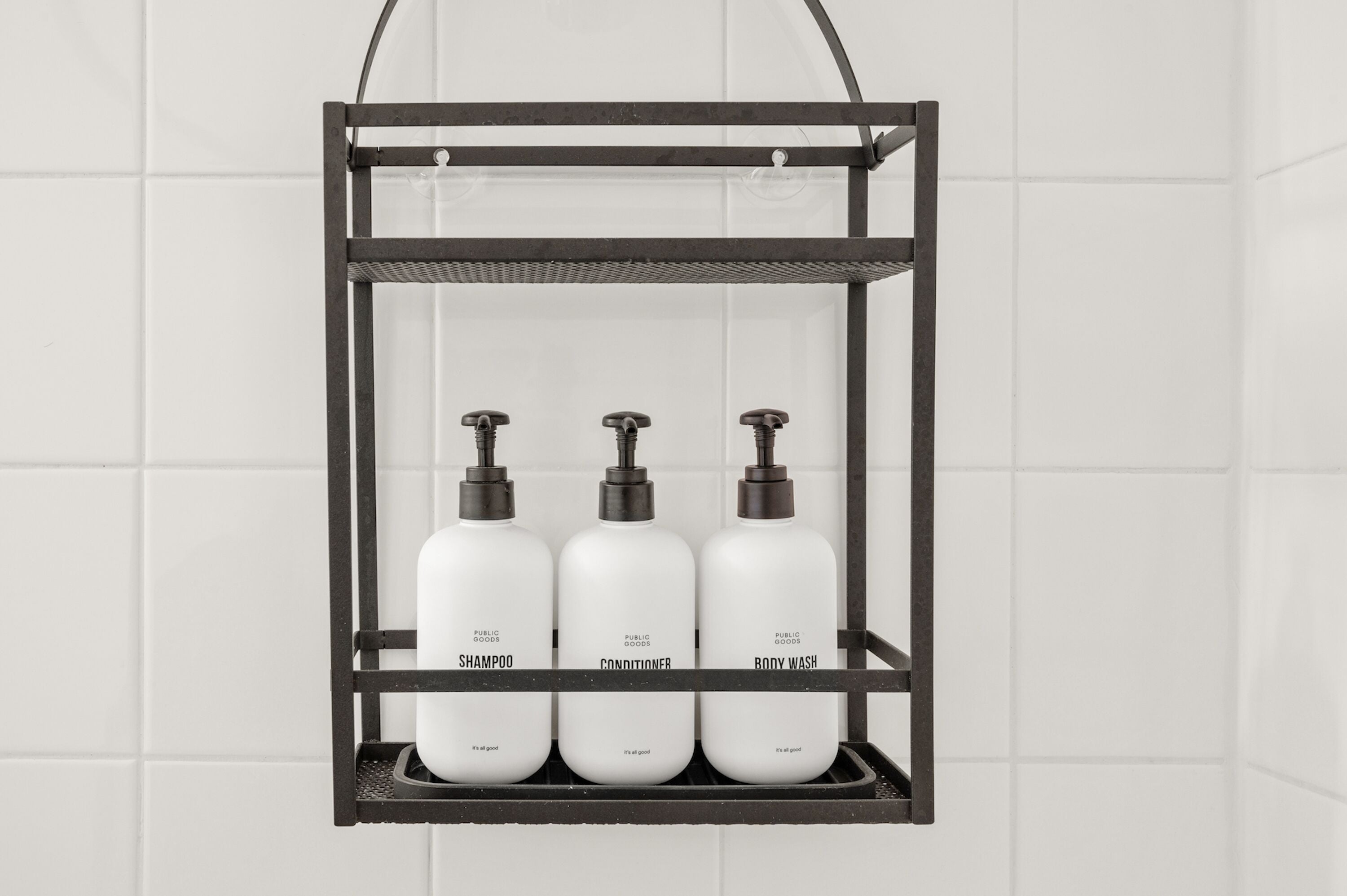 A black metal shelf with three white bottles labeled "Shampoo," "Conditioner," and "Body Wash" hanging on a tiled wall.