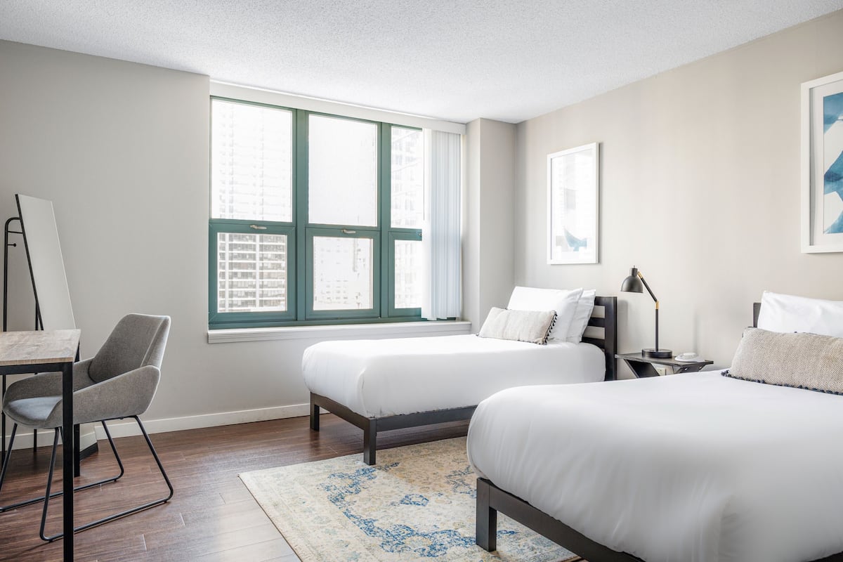 Une chambre d’hôtel avec deux lits, un bureau, une chaise et une grande fenêtre donnant sur des immeubles.