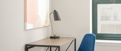 Una habitación con escritorio, una silla azul, una lámpara de pie y una ventana con persianas.